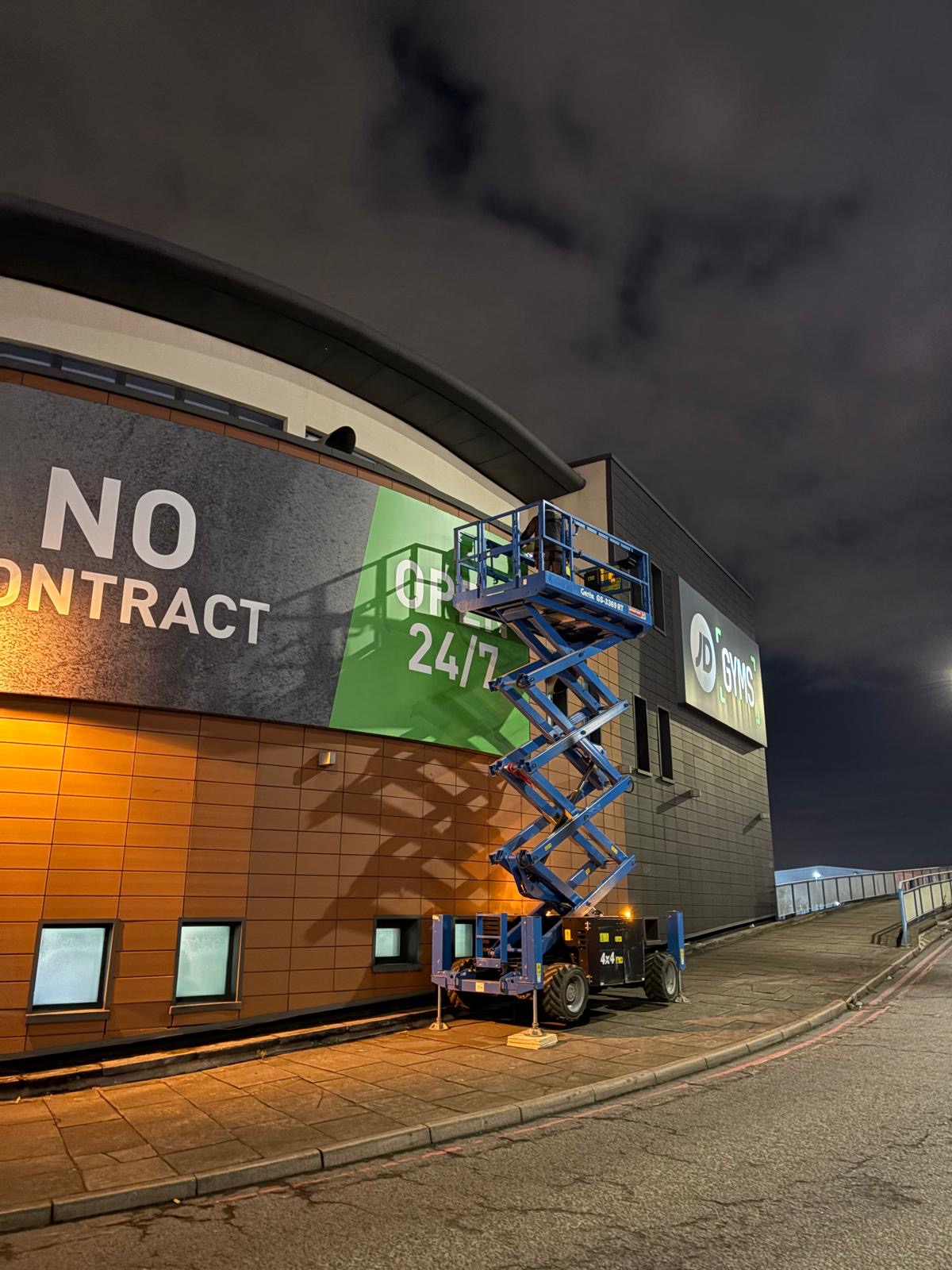 Night-time installation of the JD Gyms flex face with scissor lift access on the curved facade